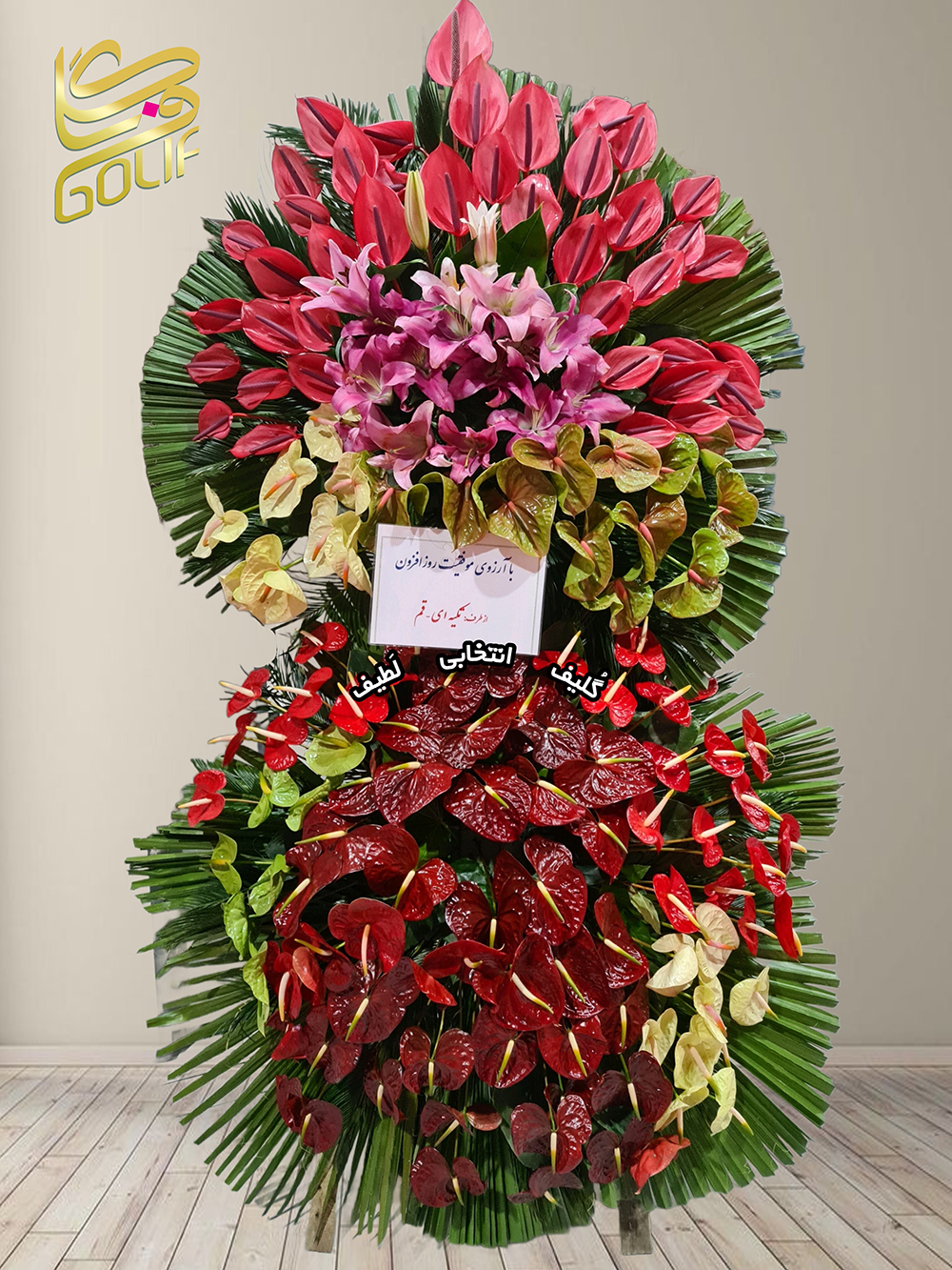 تاج گل تبریک دو طبقه (آنتوریوم، لیلیوم) گل‌های اصلی و غالب تاج گل شامل موارد زیر هستند: - **آنتوریوم قرمز، صورتی و زرد:** گل‌های برجسته در بخش‌های مختلف تاج که مرکز توجه هستند. - **لیلیوم صورتی:** در طراحی زیبای تاج به‌عنوان گل مکمل حضور دارد. ترکیب این گل‌ها به همراه برگ‌های نخل و کارت تبریک، طراحی چشم‌نوازی ایجاد کرده است. تاج گل افتتاحیه، تاج گل نمایشگاهی، تاج گل عقد و عروسی سفارش آنلاین تاج گل